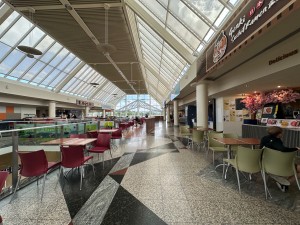 From top of lift/escalator looking straight. Path ahead. Tables and chairs along left and right. Opening to right, counter along right wall.